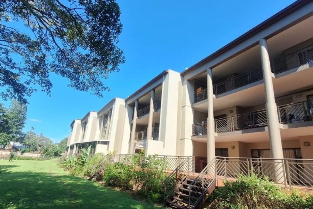 Rear view of a multi-story apartment building featuring private balconies with metal railings, concrete columns, and access to a well-maintained communal garden area.