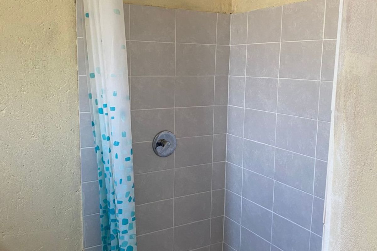 A basic shower enclosure featuring grey wall tiles, a floor-mounted drain, and a simple shower head, requiring cosmetic updates to the surrounding wall finishes.