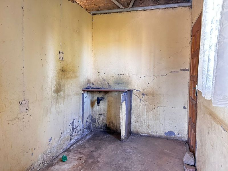 A dilapidated kitchen space requiring significant renovation, featuring exposed brick ceiling structure, damaged walls, and a basic built-in sink base.