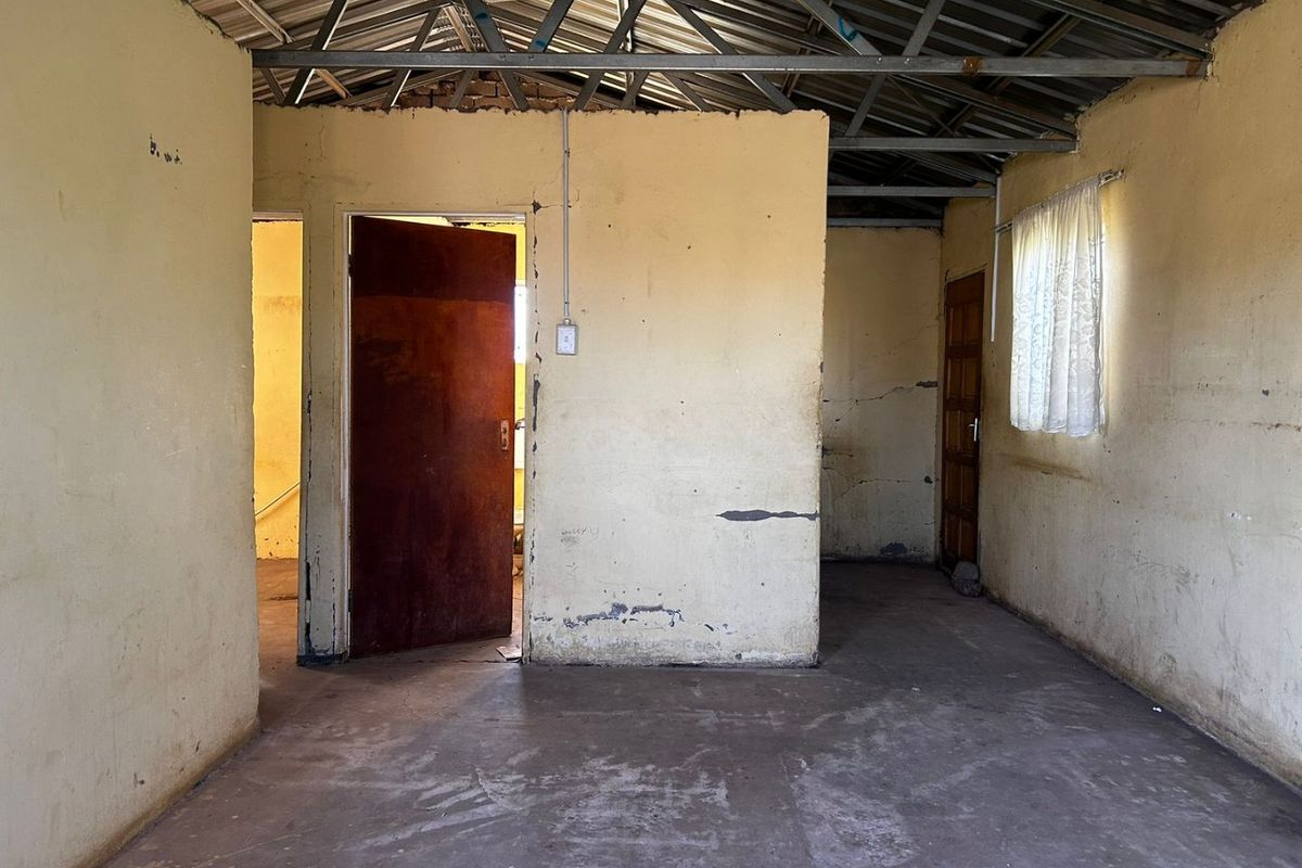 An empty, unfinished living area featuring exposed steel roof trusses, concrete flooring, and basic timber doors, requiring significant renovation and cosmetic updates.