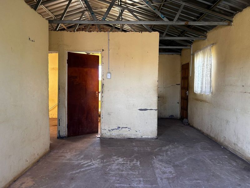 An empty, unfinished living area featuring exposed steel roof trusses, concrete flooring, and basic timber doors, requiring significant renovation and cosmetic updates.