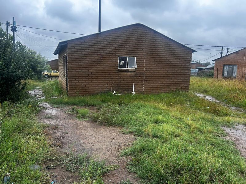 Rear view of a brick house showing signs of structural cracking, broken window glazing, and exposed exterior plumbing in an overgrown yard.