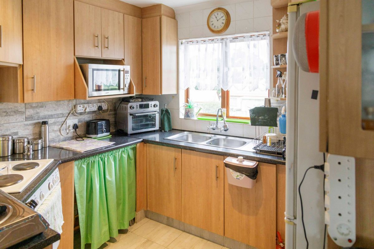 A functional kitchen featuring light wood-tone cabinetry, laminate countertops, a double stainless steel sink, and a tiled splashback with a window for natural light.