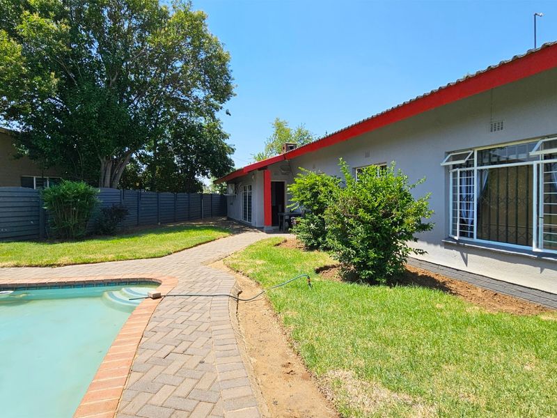 Rear exterior view featuring a swimming pool, paved walkway, and landscaped garden area alongside the main house structure.