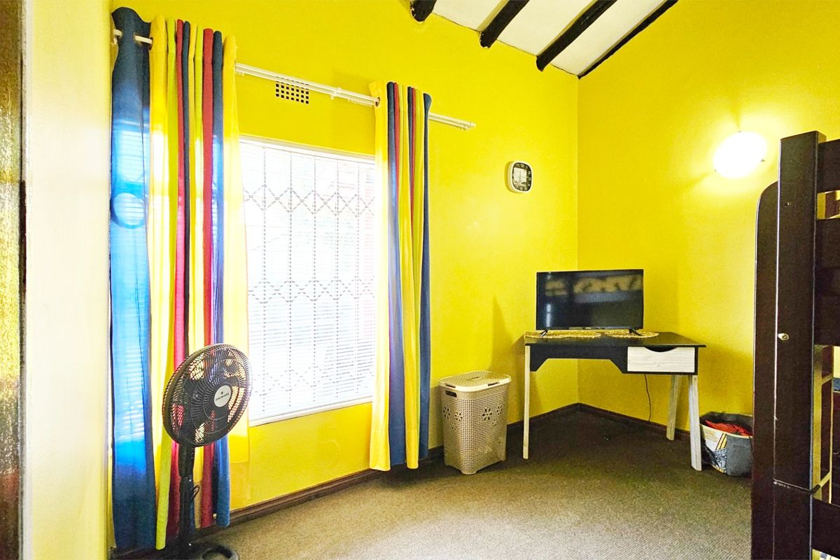 A bedroom featuring bright yellow walls, exposed ceiling beams, and a window fitted with security bars.