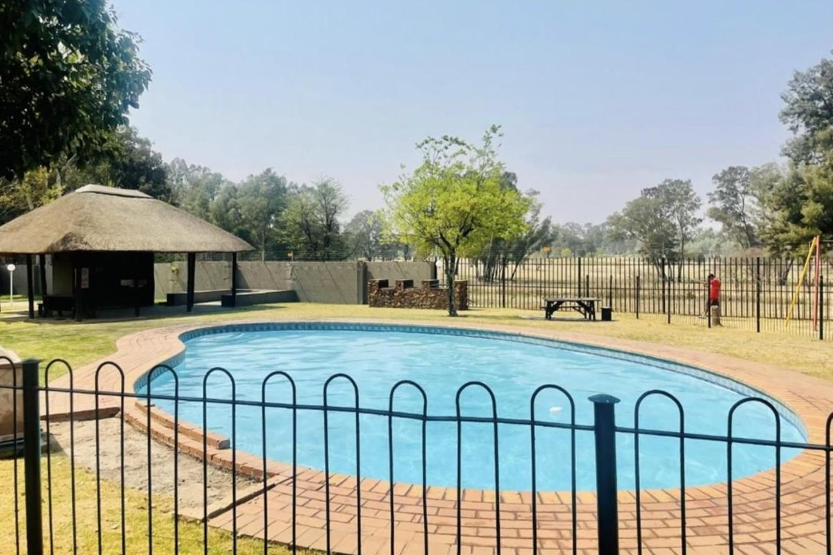 Outdoor pool area featuring a swimming pool, a thatched lapa for shade, and a stone-built braai facility within a fenced garden setting.