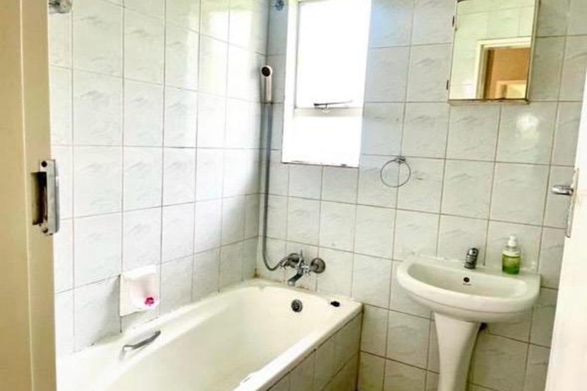 A dated bathroom featuring a standard bathtub, pedestal basin, and wall-mounted shower mixer, finished with original wall tiling.