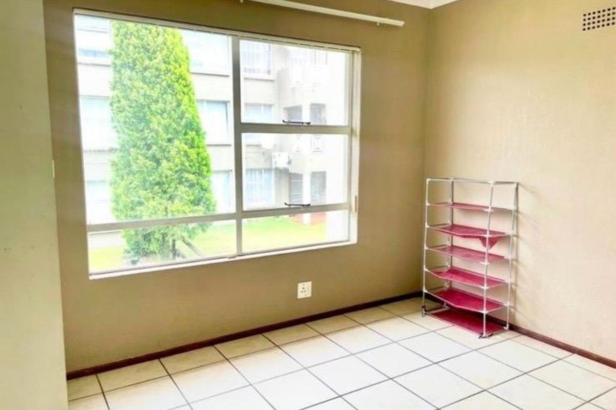 A bright, empty bedroom featuring neutral wall paint, tiled flooring, and a large window providing natural light and views of the complex.