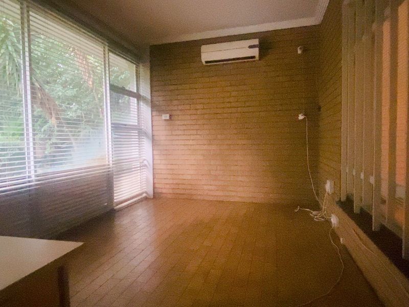A bright, empty living space featuring timber flooring, a textured brick wall, and a split system air conditioner, with large windows overlooking the garden.