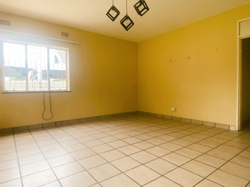 A spacious, empty living room featuring neutral floor tiles, a large window with security bars, and a simple ceiling light fixture.