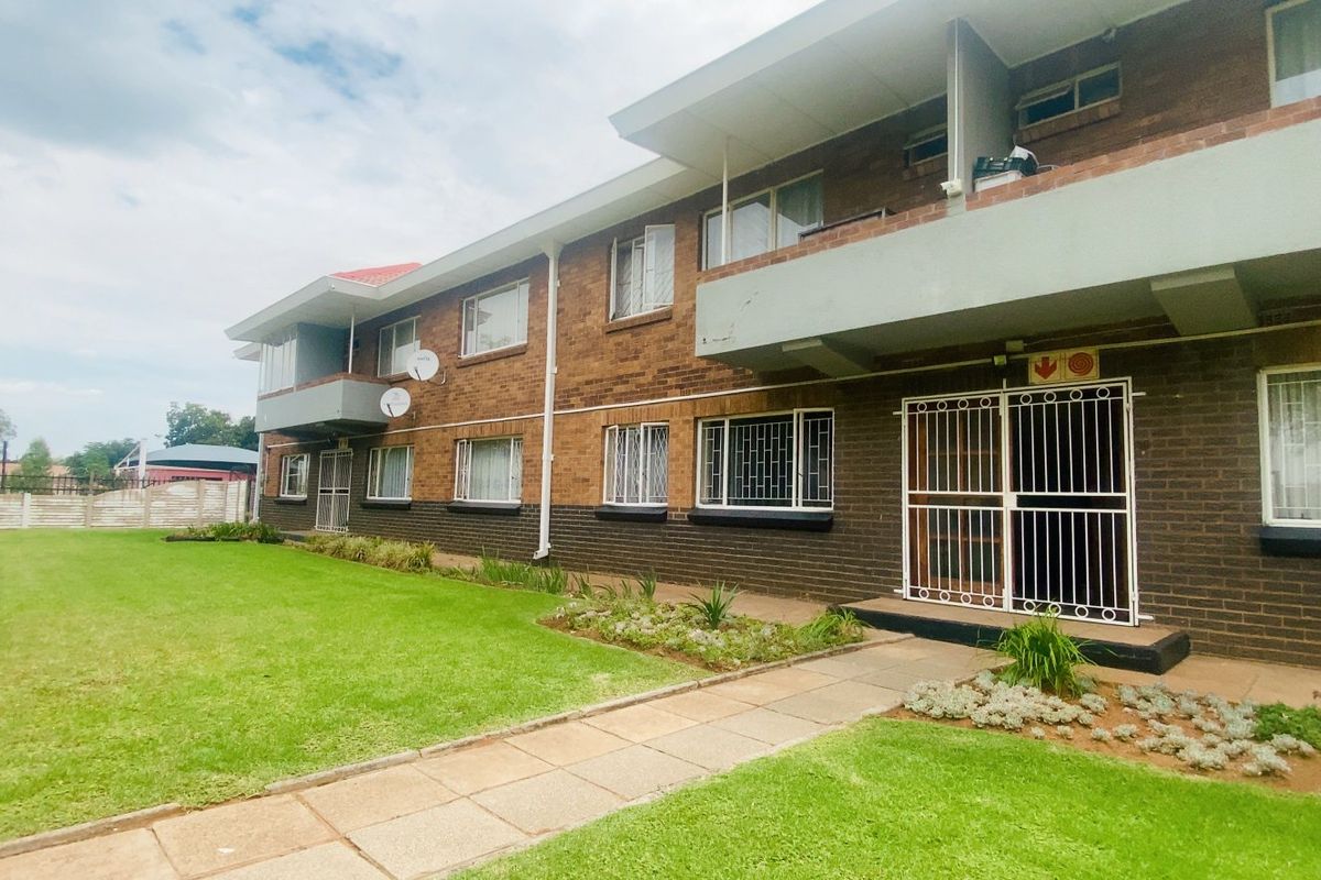 Exterior view of a multi-story apartment building featuring brick facade, private balconies, security gates, and a well-maintained communal lawn area.