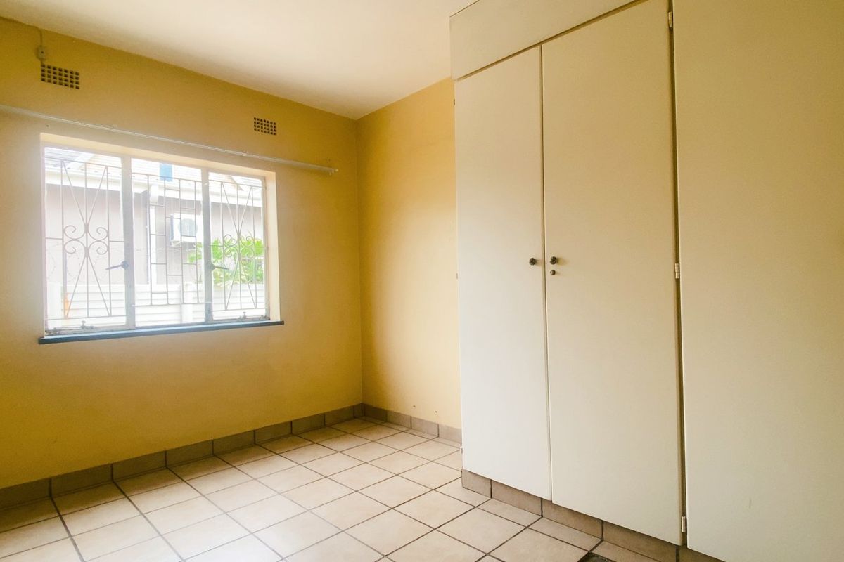 A bright, empty bedroom featuring tiled flooring, built-in cupboards, and a window with security bars.