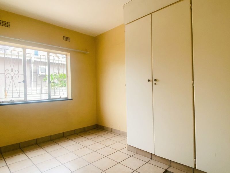 A bright, empty bedroom featuring tiled flooring, built-in cupboards, and a window with security bars.