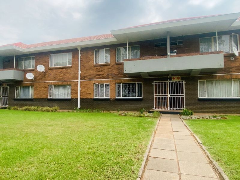 A view of the apartment complex exterior featuring a brick facade, tiled roof, and a shared front lawn with a paved walkway.