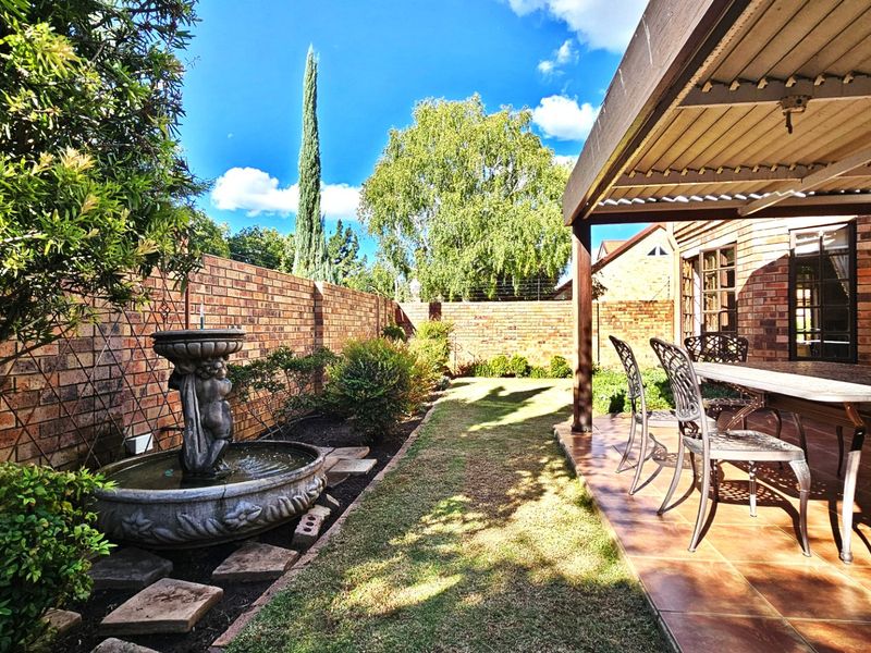 A covered patio area featuring tiled flooring, overlooking a landscaped garden with a brick boundary wall and a decorative stone water fountain.