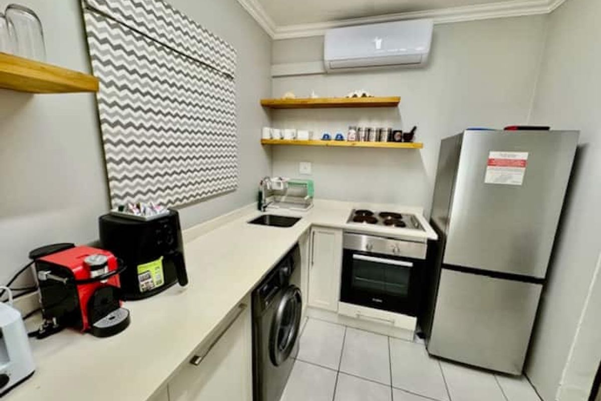 Compact kitchen featuring built-in oven, electric hob, undermount sink, floating timber shelving, and a wall-mounted air conditioning unit.