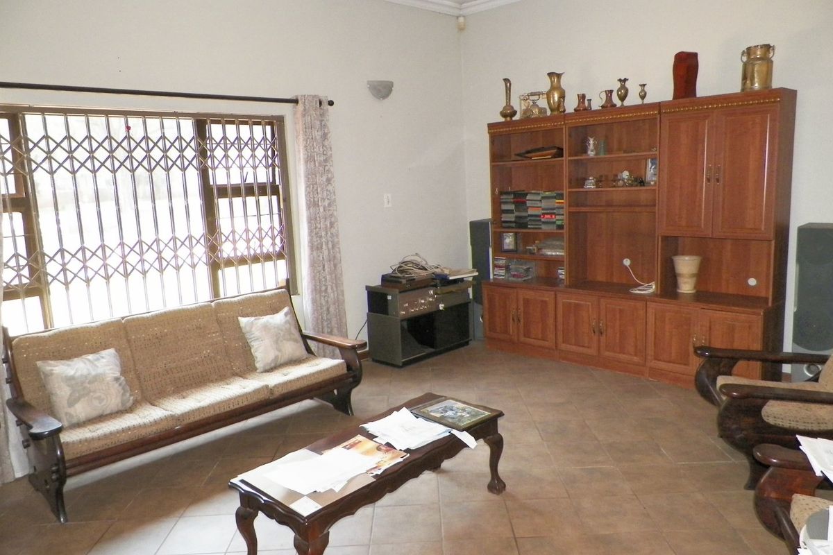 A spacious living room featuring tiled flooring, a large window with a security gate, and a built-in wooden wall unit.