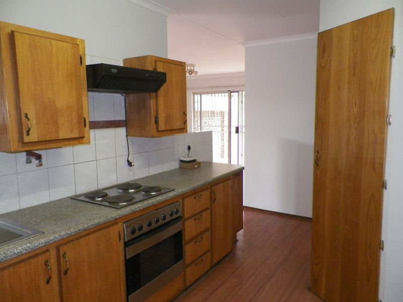 A functional kitchen featuring wooden cabinetry, laminate countertops, built-in oven and hob, and tiled splashbacks, leading into an open-plan living area.