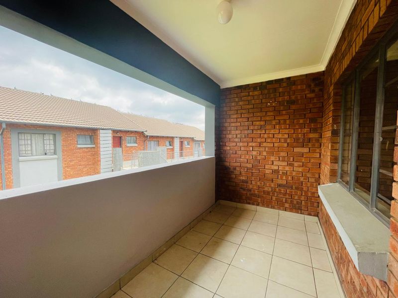 A covered balcony featuring exposed brickwork, tiled flooring, and a concrete balustrade, offering an open-air space with views of the surrounding complex.