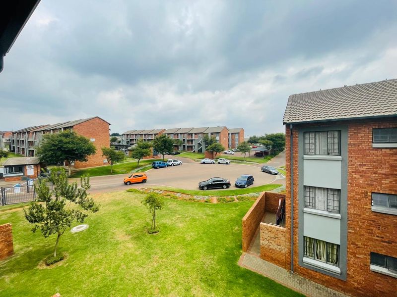 An elevated view overlooking the apartment complex grounds, featuring landscaped gardens, brick building facades, and a paved parking area.