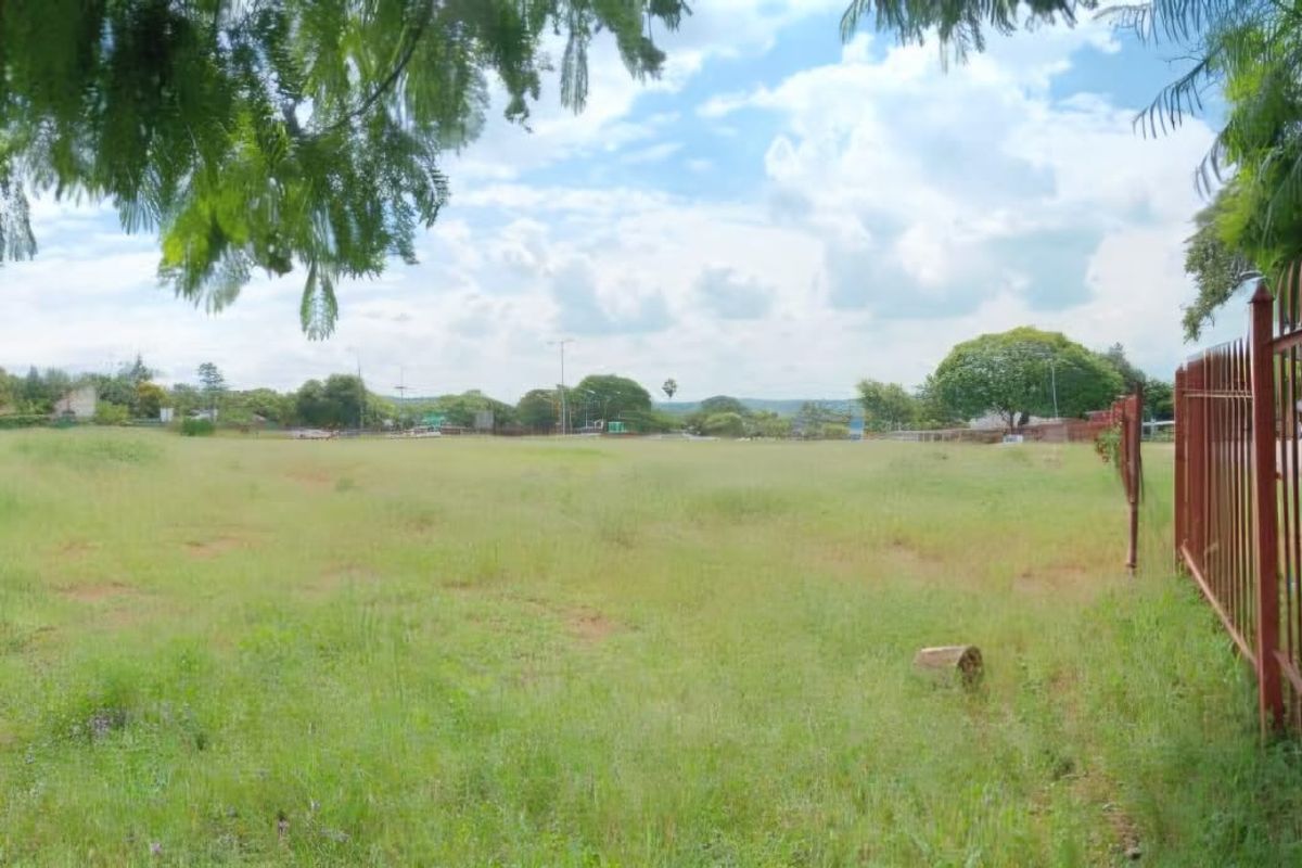 A vacant residential plot featuring open grass land and secure metal perimeter fencing, offering a clear view of the surrounding neighborhood.