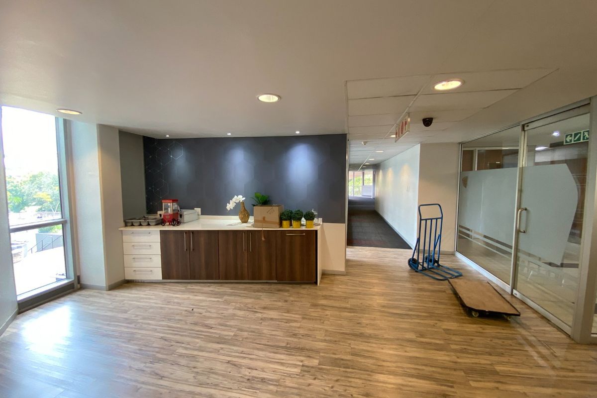 A commercial kitchenette area featuring built-in cabinetry, wood-look laminate flooring, and recessed lighting, adjacent to a glass-partitioned office corridor.