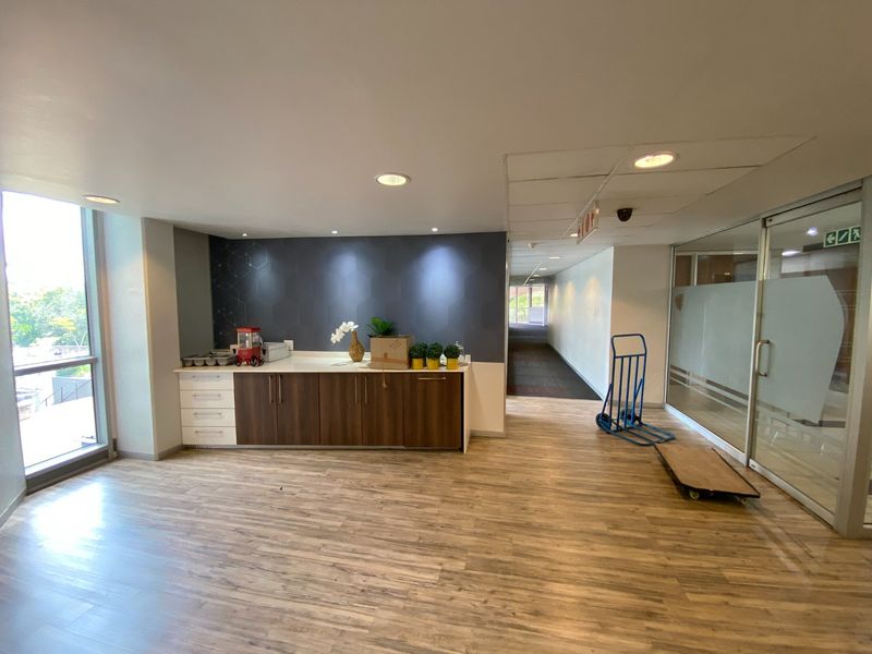 A commercial kitchenette area featuring built-in cabinetry, wood-look laminate flooring, and recessed lighting, adjacent to a glass-partitioned office corridor.
