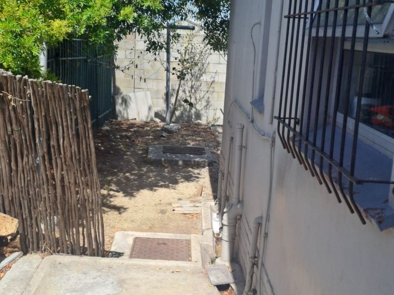 A narrow rear exterior side passage featuring concrete paving, security bars on windows, and basic boundary walling.
