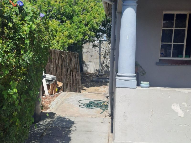 A view of the rear exterior of the house showing a concrete pathway, structural column, and garden area with visible wear to the exterior finishes.