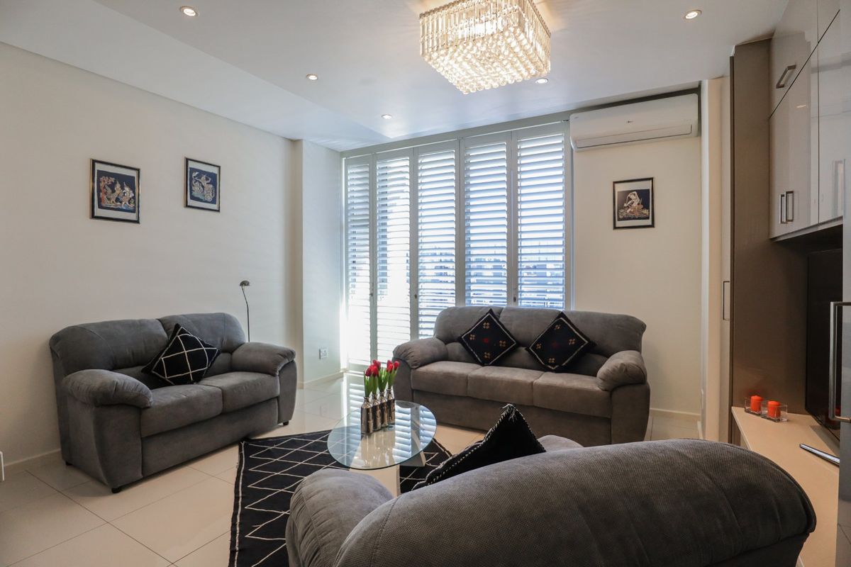 A well-maintained living room featuring tiled floors, recessed lighting, a crystal chandelier, and white plantation shutters with an integrated air conditioning unit.