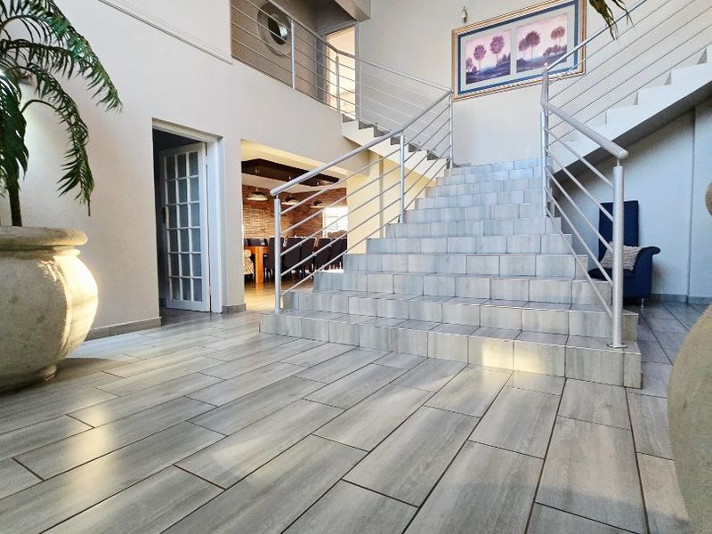 Spacious double-volume entrance hall featuring a central tiled staircase with modern stainless steel balustrades and contemporary wood-look floor tiles.