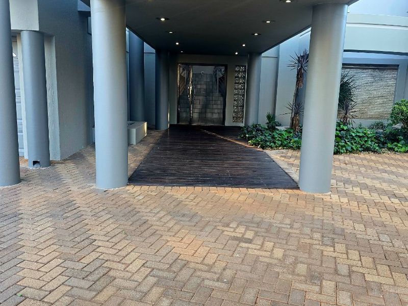 A modern covered entrance featuring a timber deck walkway, concrete columns, recessed lighting, and a decorative glass block wall with an adjacent water feature.