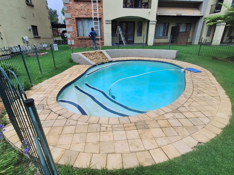 A communal garden area featuring a swimming pool with a paved surround, water feature, and metal perimeter fencing.