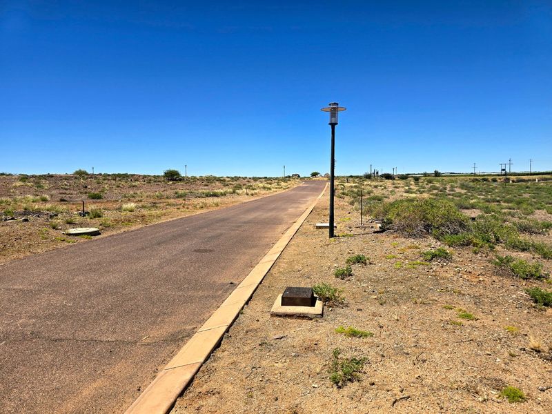 A view of a paved residential street within a vacant land development, featuring established curbing, street lighting, and utility access points.