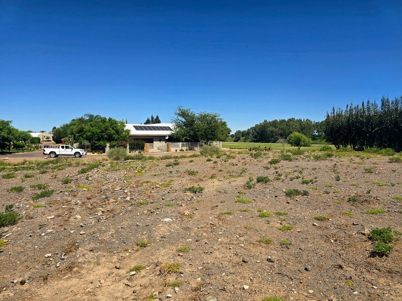 A view of a vacant residential plot of land with natural ground cover, situated in a developed neighborhood setting.