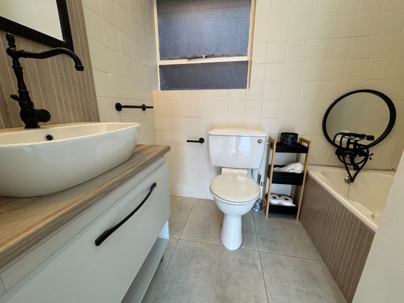 A functional bathroom featuring a modern vessel sink, vanity cabinet, toilet, and bathtub with tiled finishes and a window for natural light.