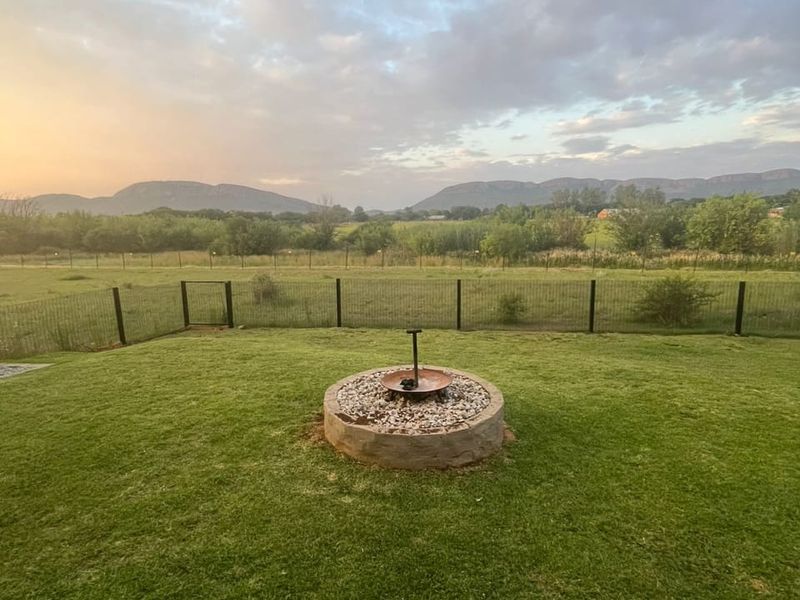 A well-maintained garden featuring a central stone fire pit, enclosed by wire fencing, offering expansive views of the surrounding landscape and mountains.