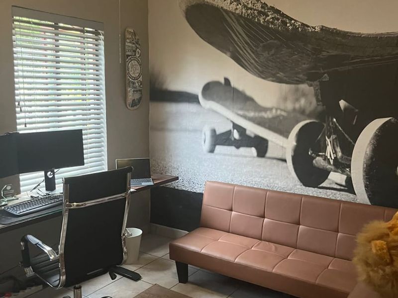 A functional home office space featuring tiled flooring and venetian blinds, currently utilized as a workspace and lounge area.