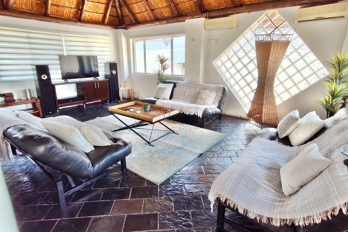 Spacious living room featuring a high thatched roof with exposed rafters, dark stone tile flooring, and a unique diamond-shaped glass block feature window.