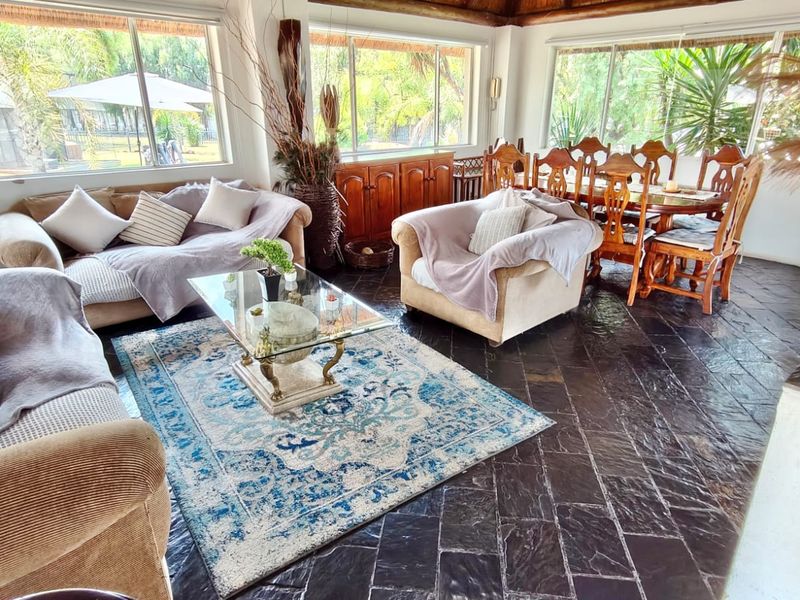 Spacious open-plan living and dining area featuring dark slate tile flooring, large windows providing natural light, and a rustic thatched roof ceiling.