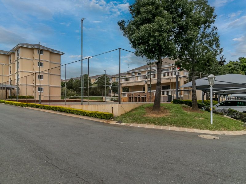Exterior view of the apartment complex featuring a fenced tennis court, paved driveway, and covered parking facilities for residents.