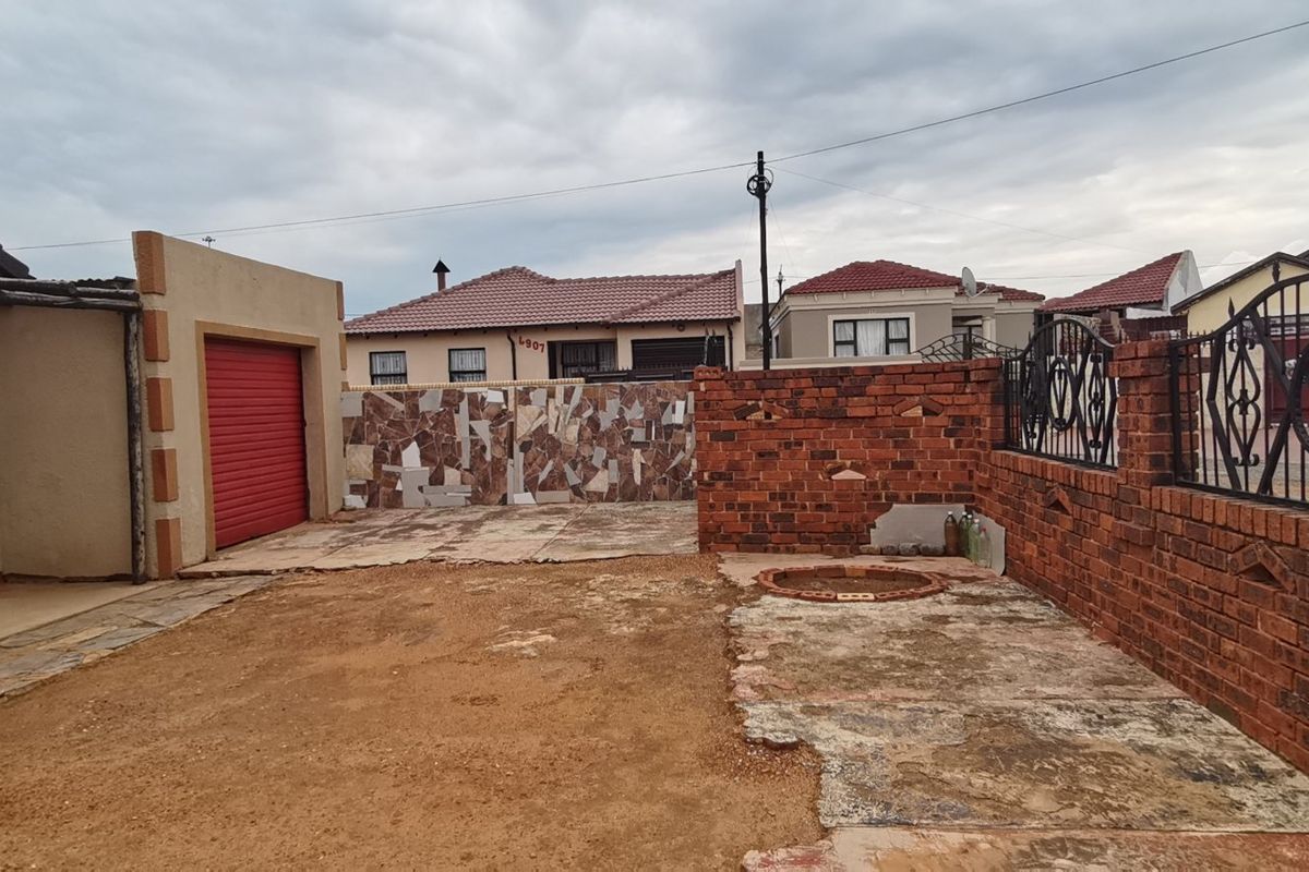 Rear exterior area featuring a brick boundary wall, metal security gate, roller shutter garage door, and a paved section requiring landscaping and surface repairs.