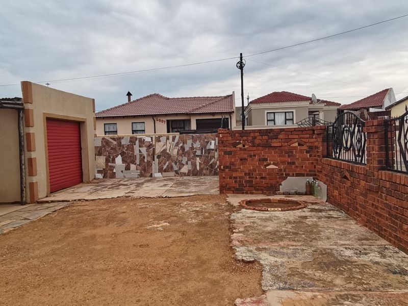 Rear exterior area featuring a brick boundary wall, metal security gate, roller shutter garage door, and a paved section requiring landscaping and surface repairs.