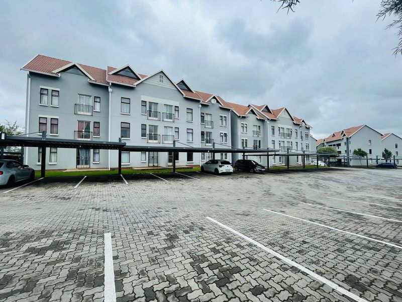 Exterior view of a multi-story apartment complex featuring a large paved parking area with covered parking bays and residential balconies.