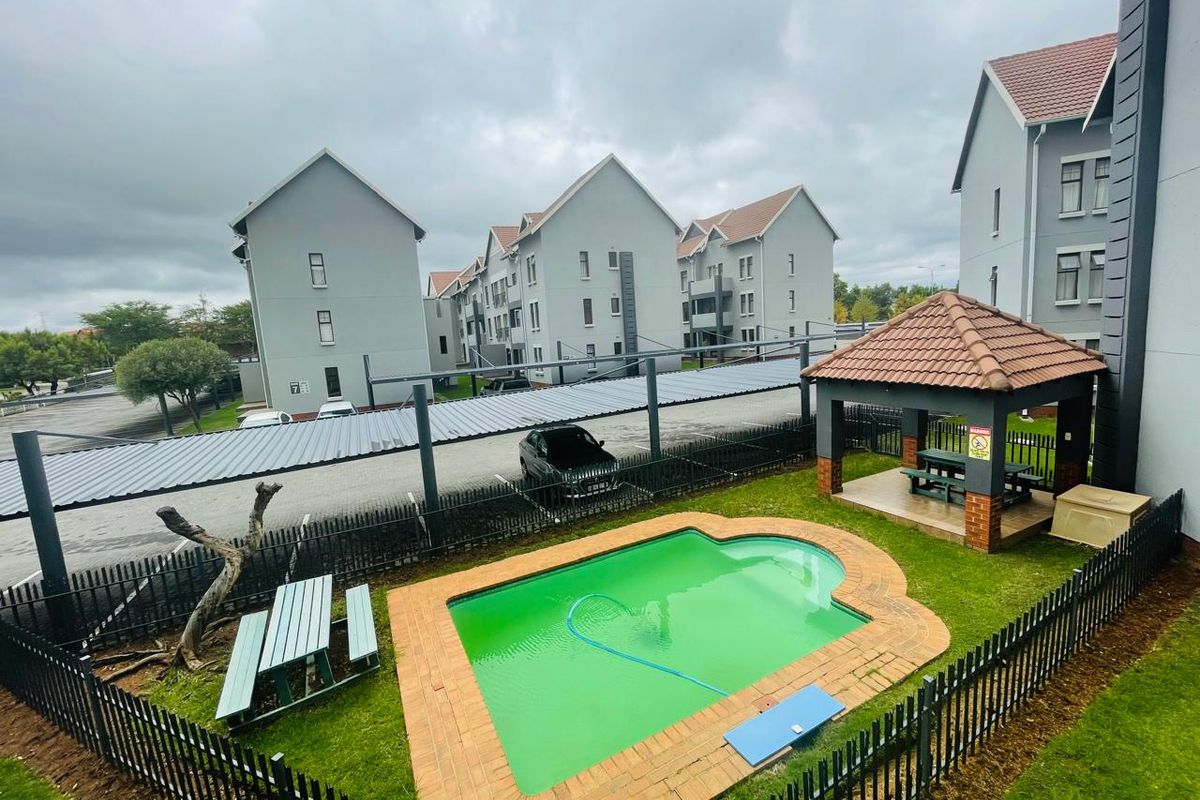 Communal pool area featuring a brick-edged swimming pool, a thatched gazebo, and metal perimeter fencing within an apartment complex setting.
