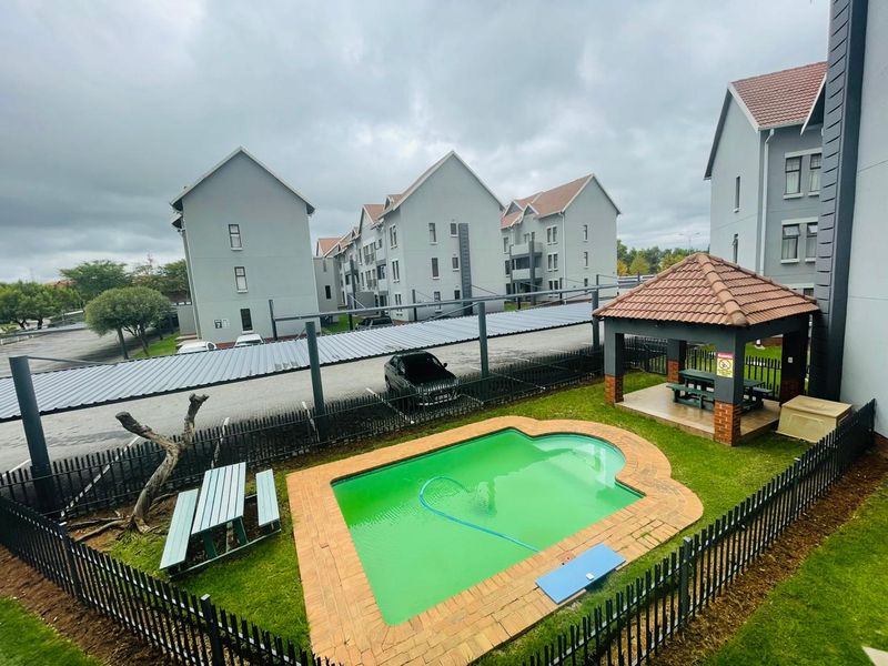 Communal pool area featuring a brick-edged swimming pool, a thatched gazebo, and metal perimeter fencing within an apartment complex setting.