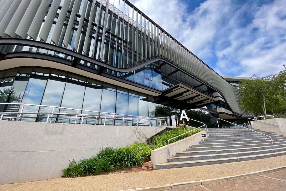 Modern commercial building facade featuring a glass curtain wall, vertical sun louvers, and a prominent entrance staircase with stainless steel handrails.