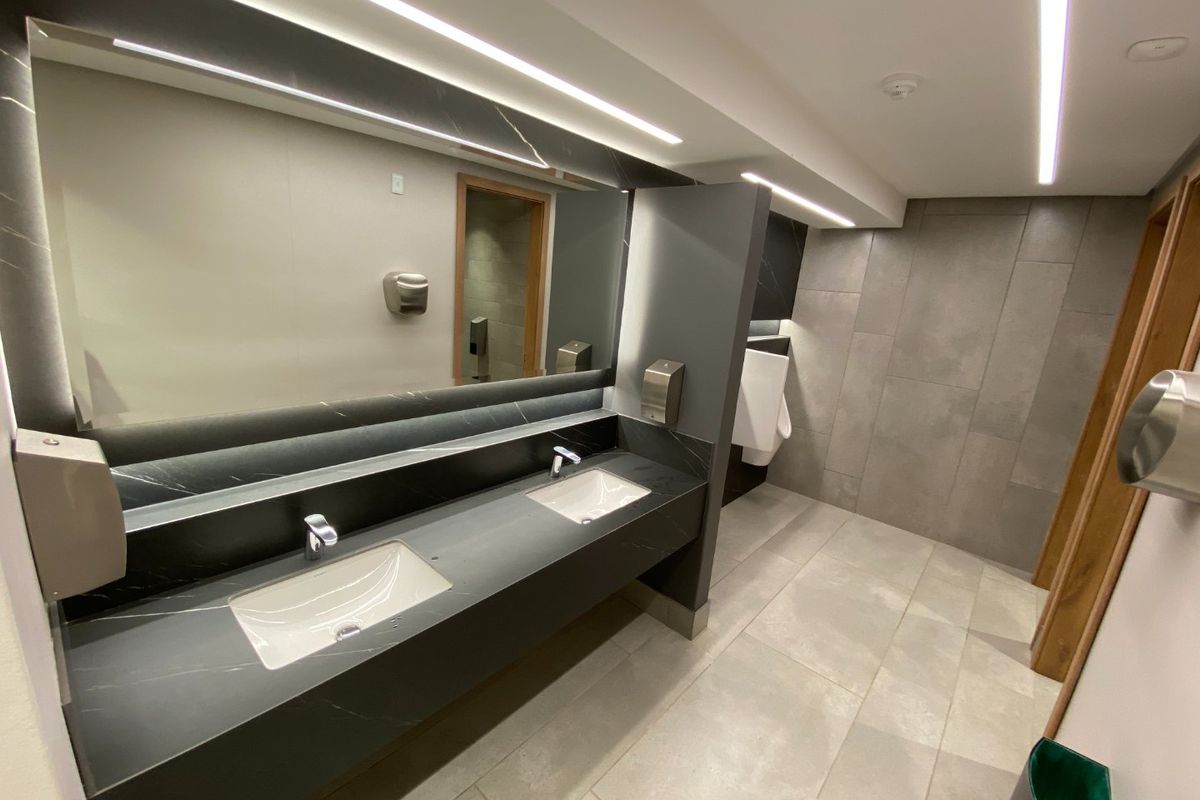 Modern commercial bathroom featuring a sleek double vanity with integrated sinks, large mirror, contemporary grey tiling, and recessed linear lighting.