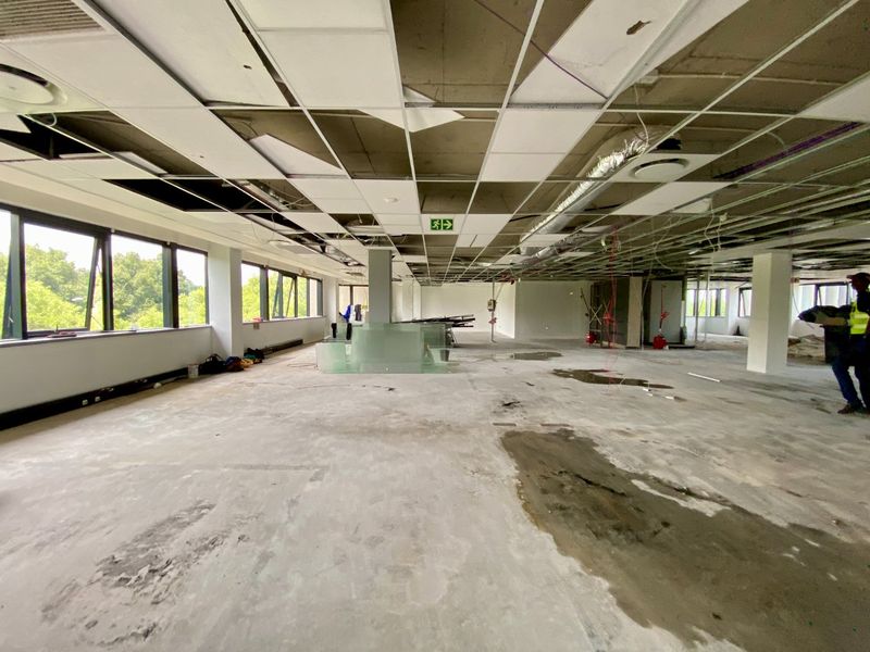 Large open-plan commercial office space currently undergoing renovation, featuring exposed ceiling infrastructure, concrete flooring, and expansive windows providing natural light.