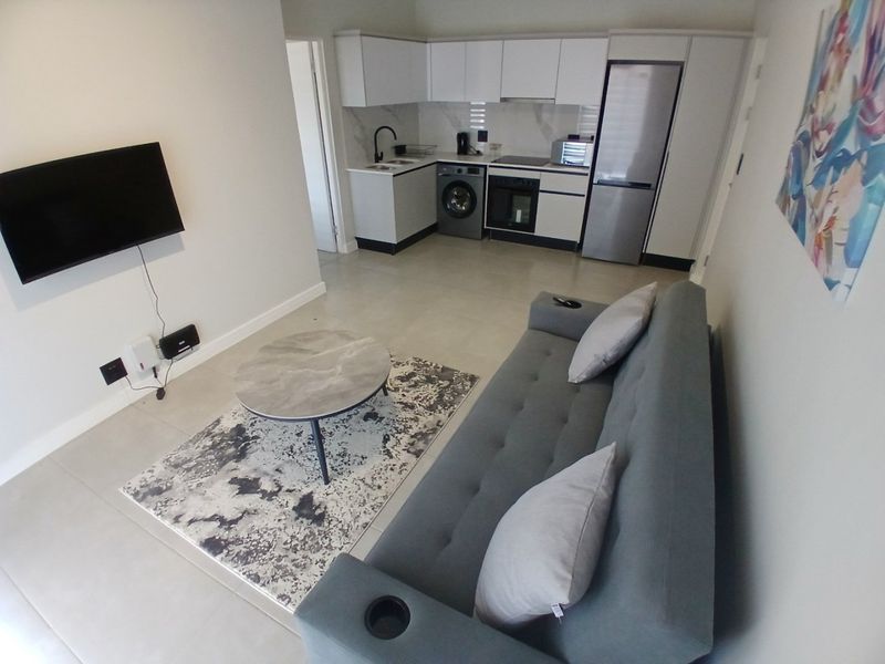 Modern open-plan living area featuring a compact kitchen with built-in cabinetry, integrated appliances, and neutral tiled flooring throughout.
